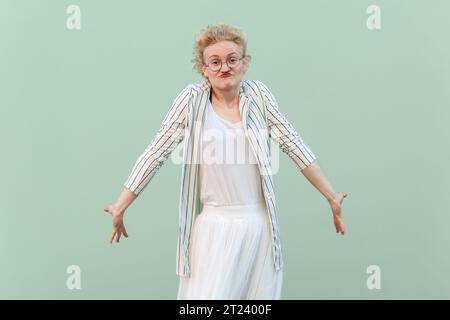 Ritratto di una donna bionda adulta confusa e perplessa che indossa camicia e gonna a righe, spalle alle spalle, non sa rispondere e cosa fare. Riprese in studio in interni isolate su sfondo verde chiaro. Foto Stock