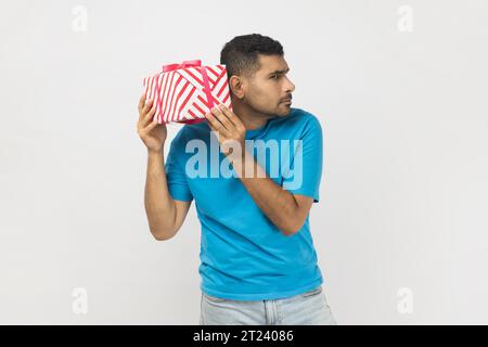 Ritratto di un uomo curioso e non rasato che indossa una T-shirt blu con un'espressione facciale interessata, scuotendo la scatola del regalo, pensando a cosa dentro. Riprese in studio in interni isolate su sfondo grigio. Foto Stock
