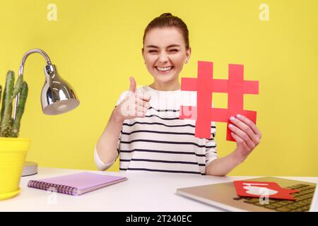 Blogger donna sorridente di buon umore, con hashtag rosso e pollice in alto come un gesto, scattare selfie POV, sedersi sul posto di lavoro. Studio interno girato isolato su sfondo giallo. Foto Stock