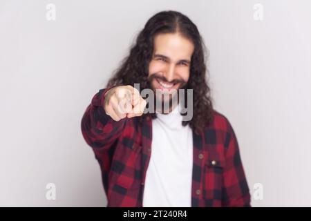 CE l'hai fatta. Uomo barbuto felice con lunghi capelli ricci in camicia rossa a scacchi puntata alla fotocamera con le dita indice, sceglie qualcuno sorride ampiamente. Riprese in studio in interni isolate su sfondo grigio. Foto Stock