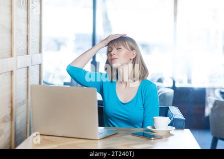 Ritratto di triste turbato disperazione giovane donna con i capelli biondi in camicia blu che lavora su un computer portatile, facendo gesto palmare, ha problemi con il lavoro. Foto in interni in un bar con una grande finestra sullo sfondo. Foto Stock