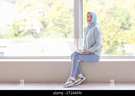 Donna musulmana elegante che utilizza un computer portatile vicino alla finestra a casa Foto Stock