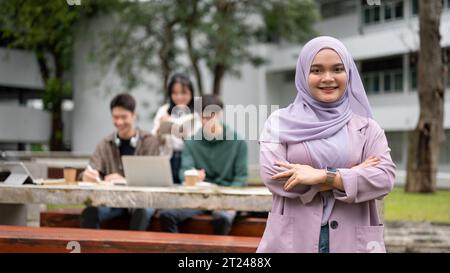 Una giovane studentessa universitaria asiatica-musulmana sorridente e affascinante in un hijab viola si trova in piedi con le braccia incrociate mentre i suoi amici lavorano in togeth Foto Stock