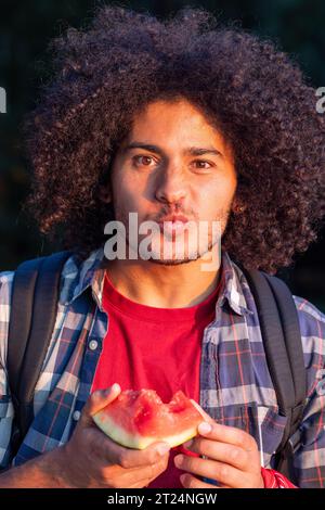 Un momento affascinante catturato come un affascinante uomo arabo millenario con lunghi capelli ricci assapora un delizioso pezzo di anguria nel caldo bagliore del sole che tramonta, un assaggio di gioia estiva. Arabian Man che si gode l'anguria al tramonto. Foto di alta qualità Foto Stock