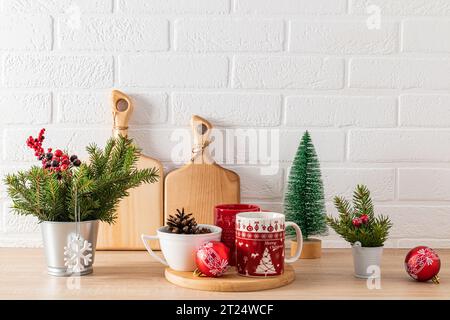 Tazze rosse con motivo natalizio su vassoio di legno tra decorazioni verdi con rami di abete sul ripiano della cucina per le vacanze. Casa accogliente Foto Stock