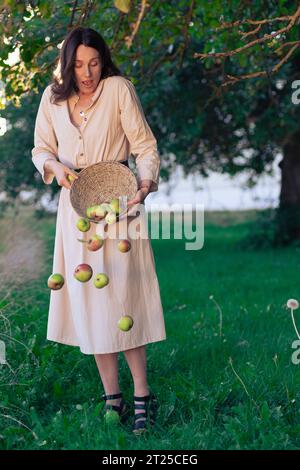 La bellissima ragazza disperde le mele e cadono dal cesto sull'erba. Giovane donna in bel vestito che posa nel frutteto di mele con la caduta Foto Stock