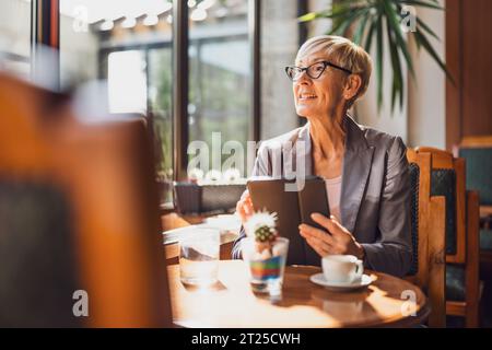 Una donna matura è seduta in una caffetteria e si sta rilassando. Sta bevendo caffè e sta usando un tablet digitale. Foto Stock