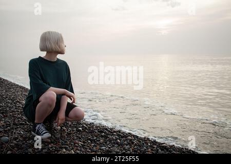 Bella giovane donna adulta con capelli biondi corti seduta sulla spiaggia di mare all'aperto Foto Stock
