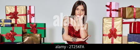 bella signora in abito rosso vicino a regali con mano sulla guancia che guarda la macchina fotografica, regali di natale, striscione Foto Stock