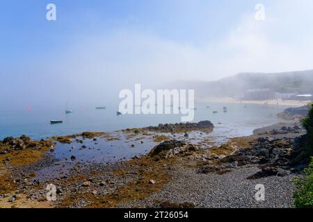 Il cielo blu inizia a sfondare attraverso una bassa riva di nebbia costiera che si trova sopra il villaggio di pescatori di Porthdinllaen. Foto Stock