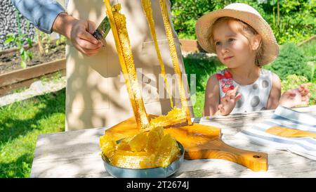 Una ragazza mangia il miele in un favo. Una ragazza con un cappello guarda suo nonno tagliare il miele fresco in un favo. Una ragazza con la voglia di dolce sta aspettando Foto Stock