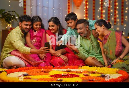 Gruppo di una famiglia mista indiana felice che vede il cellulare durante la festa della famiglia diwali davanti ai rangoli di fiori - concetto di emozione Foto Stock