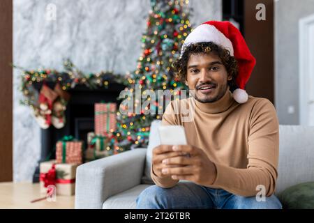 Felice uomo di natale sorridente in cappello rosso di babbo natale seduto sul divano in salotto a casa, uomo ispanico che tiene il telefono e guarda la fotocamera, utilizzando l'app per smartphone. Foto Stock