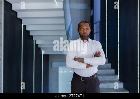 Ritratto di un uomo afro-americano con una camicia bianca in piedi in un centro di un ufficio e incrocia le braccia sul petto, guardando con sicurezza e serietà la fotocamera. Foto Stock