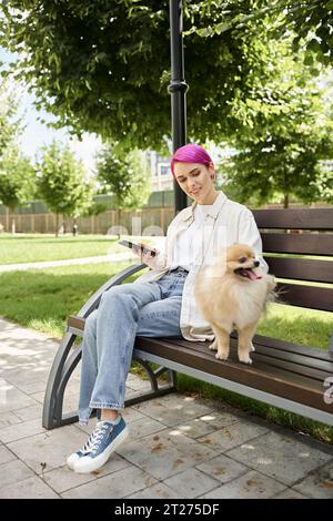 donna sorridente dai capelli viola con smartphone seduto sulla panchina nel parco vicino al divertente pomeranian spitz Foto Stock