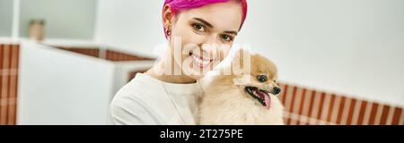 gioiosa dog sitter dai capelli viola con simpatico pomeranian spitz che guarda la macchina fotografica in un hotel per animali domestici, banner Foto Stock