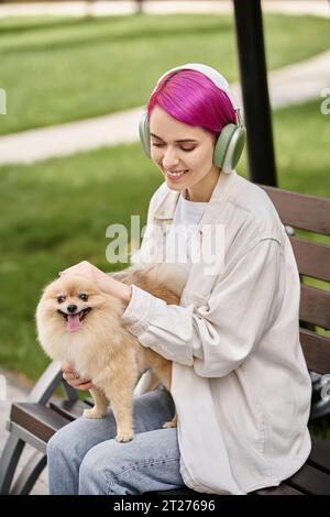 donna felice che ascolta musica in cuffia e che accarezza l'adorabile pomeranian spitz sulla panchina nel parco Foto Stock