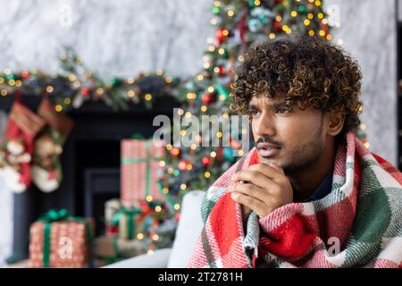 Uomo congelante seduto sotto una coperta a casa sul divano del soggiorno, il riscaldamento è spento, il riscaldamento dell'appartamento non funziona, gente ispanica per Natale vicino all'albero. Foto Stock
