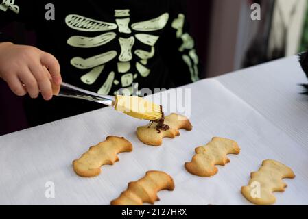 Il ragazzo decora biscotti di pan di zenzero di halloween su piatti con glassa di cioccolato. Prelibatezze culinarie per festeggiare halloween. Stile di vita Foto Stock