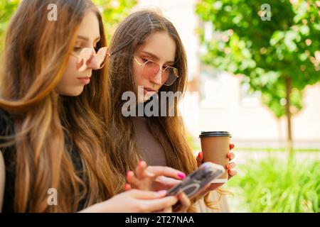 Sorelle gemelle stupite con occhiali da sole che utilizzano i social media con uno smartphone seduto su una panchina in città e che tiene all'aperto una tazza di caffè di carta usa e getta Foto Stock