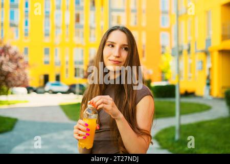 Una giovane donna carina che apre una bottiglia di succo di limone di plastica arancione in piedi sulla strada della città, sullo sfondo di edifici colorati Foto Stock