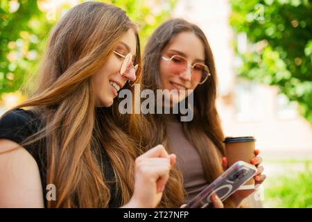 Sorelle gemelle stupite con occhiali da sole che utilizzano i social media con uno smartphone seduto su una panchina in città e che tiene all'aperto una tazza di caffè di carta usa e getta Foto Stock