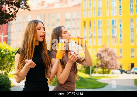 Giovani sorelle gemelle urbane con occhiali da sole freschi che aprono una bottiglia di succo di limone sullo sfondo della strada della città Foto Stock