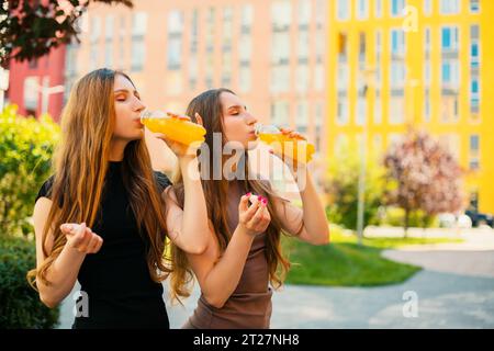 Giovani sorelle gemelle urbane con occhiali da sole freschi che aprono una bottiglia di succo di limone sullo sfondo della strada della città Foto Stock