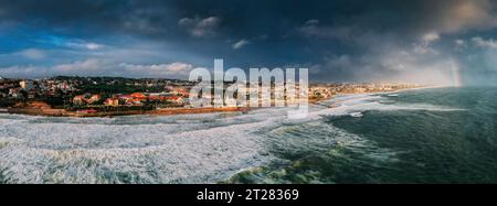 Vista panoramica aerea della costa di Cascais con un arcobaleno che si riflette sui mari tempestosi al largo della costa all'estrema destra Foto Stock