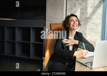 Ritratto di una donna d'affari in completo, che lavora dall'ufficio, che ride mentre fa una chiacchierata su un video, guarda un notebook, parla online Foto Stock