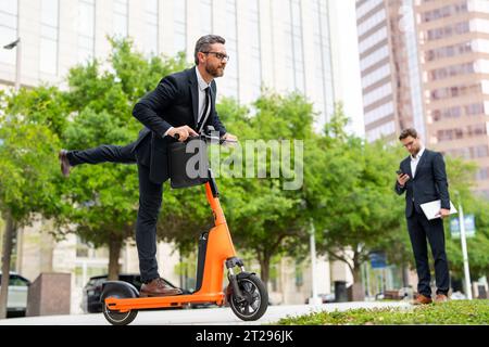 Un uomo d'affari eccitato e divertente su uno scooter elettrico per le strade cittadine. Impiegato uomo d'affari in tuta avvocato aziendale in tuta che guida e-scooter. Uomo d'affari Foto Stock