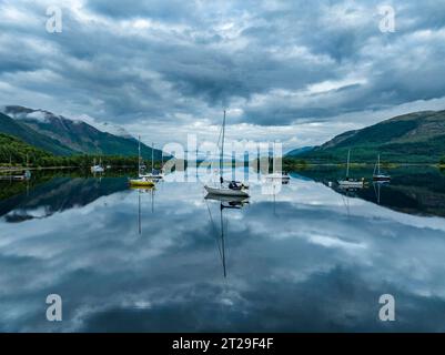 Vista aerea, atmosfera mattutina con riflessi d'acqua sul lago di loch Leven di acqua dolce, con barche a vela parcheggiate sulla riva del villaggio di Glen Coe Foto Stock