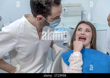 Una ragazza dall'aspetto preoccupata sulla sedia del dentista davanti al dottore si lamenta per il suo dolore gengivale e per aver tenuto la mano sulla guancia. Foto Stock