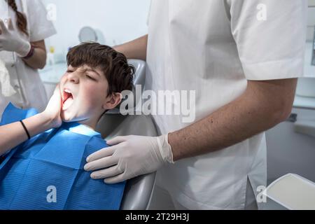 Un ragazzo preoccupato sulla sedia del dentista di fronte al dottore si lamenta a causa del suo dolore gengivale e tenendo la mano sulla guancia. Foto Stock