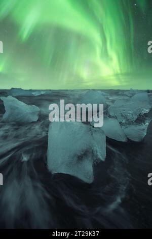 Pittoresco scenario di acqua di mare che si tuffa con pezzi di ghiaccio spezzato nella spiaggia di sabbia nera contro il cielo verde dell'aurora boreale a Diamond Beach in Islanda Foto Stock