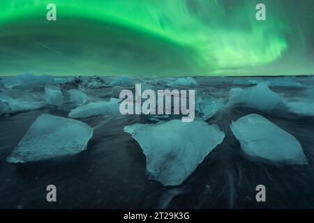 Pittoresco scenario di acqua di mare che si tuffa con pezzi di ghiaccio spezzato nella spiaggia di sabbia nera contro il cielo verde dell'aurora boreale a Diamond Beach in Islanda Foto Stock