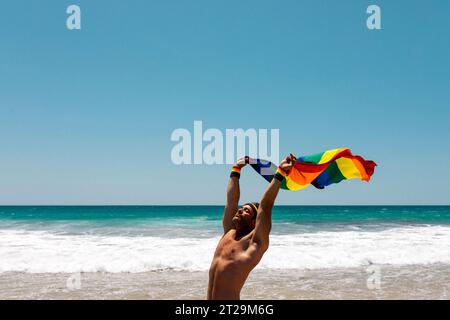 Foto con vista laterale di un uomo orgoglioso che regge una bandiera arcobaleno LGBT con gli occhi chiusi dal mare ondulato sulla spiaggia Foto Stock