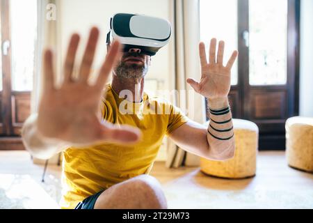 Il maschio barbuto in maschera VR si allunga alle braccia mentre si allena in un appartamento leggero Foto Stock