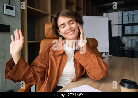 Imprenditrice donna, direttrice d'ufficio seduta nella sala conferenze, ascoltando musica con auricolari wireless e ballando, ridendo e sorridendo, divertendosi Foto Stock