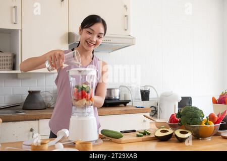 La giovane donna asiatica sana e bella con abiti informali è sorridere mettendo cetriolo, banana e frutta di pomodoro nel frullatore durante la preparazione del frullato mattutino Foto Stock