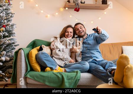 Una giovane coppia felice e il loro piccolo cane si siedono sul divano a casa e guardano la TV durante le vacanze di Natale. Foto Stock