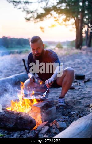 Un brutale uomo con una barba cucina il cibo in una padella su un fuoco in natura. Pancetta da cucina sulla riva del fiume. Campeggio, estate Foto Stock