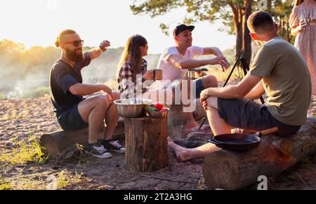 La compagnia di ragazze e ragazzi festeggia una vacanza nella natura. Stringono i bicchieri con le bevande. Riposatevi in estate nella foresta sulle rive della riva del fiume Foto Stock