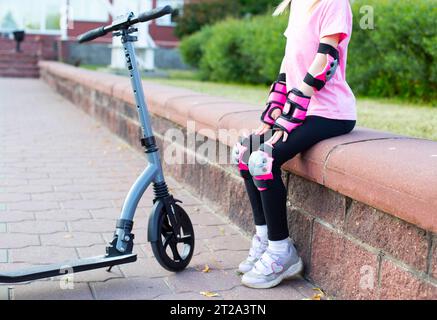 Una ragazza con un equipaggiamento protettivo rosa siede su un marciapiede accanto a uno scooter. Il concetto di sicurezza durante la guida di uno scooter. Foto Stock