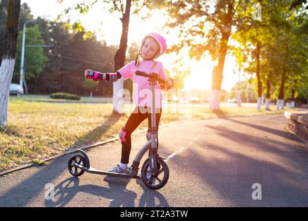 Una bella bambina di sette anni con un casco rosa e un equipaggiamento protettivo si pone con uno scooter in città sullo sfondo del tramonto. Foto Stock