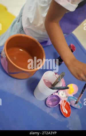 Un bambino che indossa un grembiule colorerà un vaso di fiori con vernice e una spazzola su un tavolo blu in classe. Foto Stock