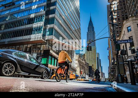 Una strada con alti edifici per uffici a New York, insieme a un'auto e un ciclista illuminati dalla luce riflessa dagli edifici, in piedi ad una r Foto Stock