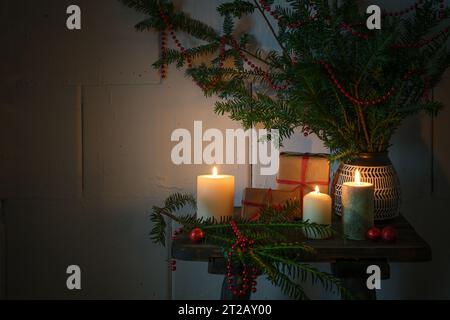 Natale con candele in fiamme, regali, rami sempreverdi e decorazioni su un piccolo tavolo di legno, luce calda e accogliente nella stagione buia, spazio fotocopie, Foto Stock