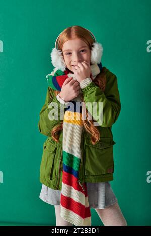 allegra ragazza con sciarpa a righe e cuffie invernali sorridente su uno sfondo turchese e comodi strati Foto Stock