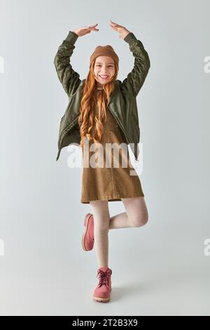 ragazza allegra in outfit invernale con stivali e cappello sorridenti e in posa da ballerina su sfondo grigio Foto Stock